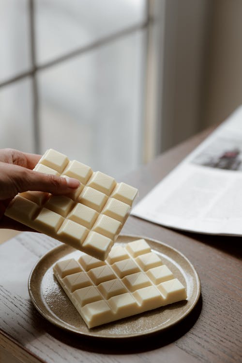 Crop unrecognizable person holding piece of white chocolate while resting at table and reading magazine