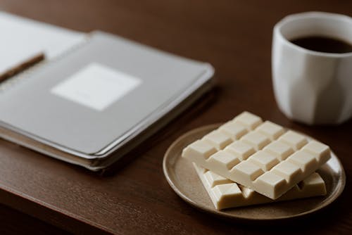 Free High angle of delicious white chocolate on ceramic plate near mug of fresh coffee and paper notebook Stock Photo