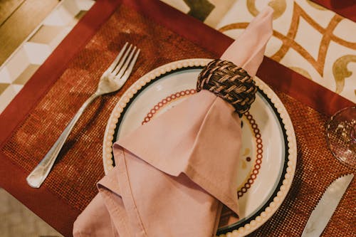 From above layout of table setting with pink napkin served on fancy ceramic plate on place mat near cutlery