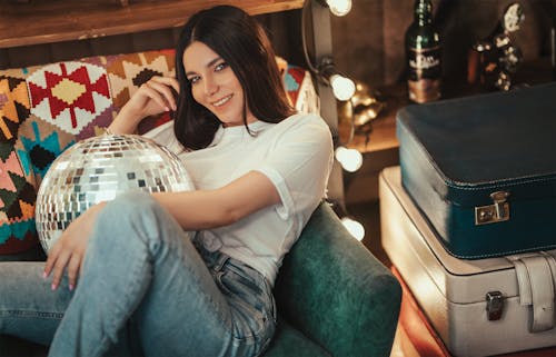 Cheerful young female with brown hair smiling and sitting on cozy couch with disco ball