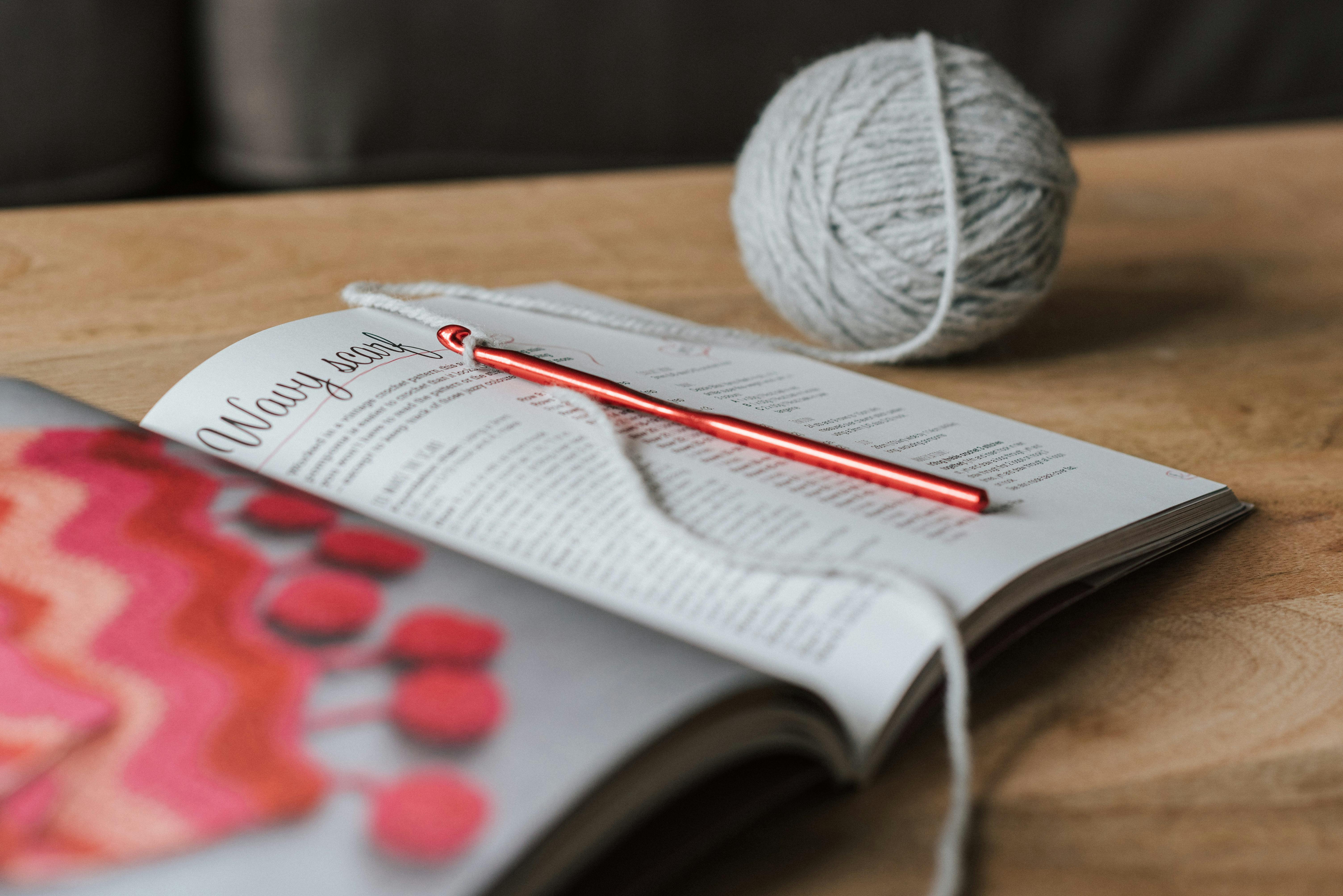 Spherical gray ball of thread and red crochet needle with magazine on beige wooden table in daytime on blurred background