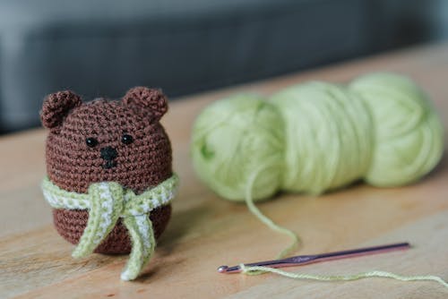 Free Closeup of small dark brown crochet toy bear and crochet next to light green threads on wooden table in bright room on blurred background Stock Photo