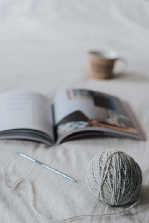 Composition of gray yarn ball and crochet needle placed on comfy bed with opened magazine and cup of hot drink