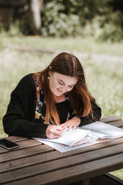 Free Young cheerful female doing homework on timber table on blurred background of nature in park Stock Photo