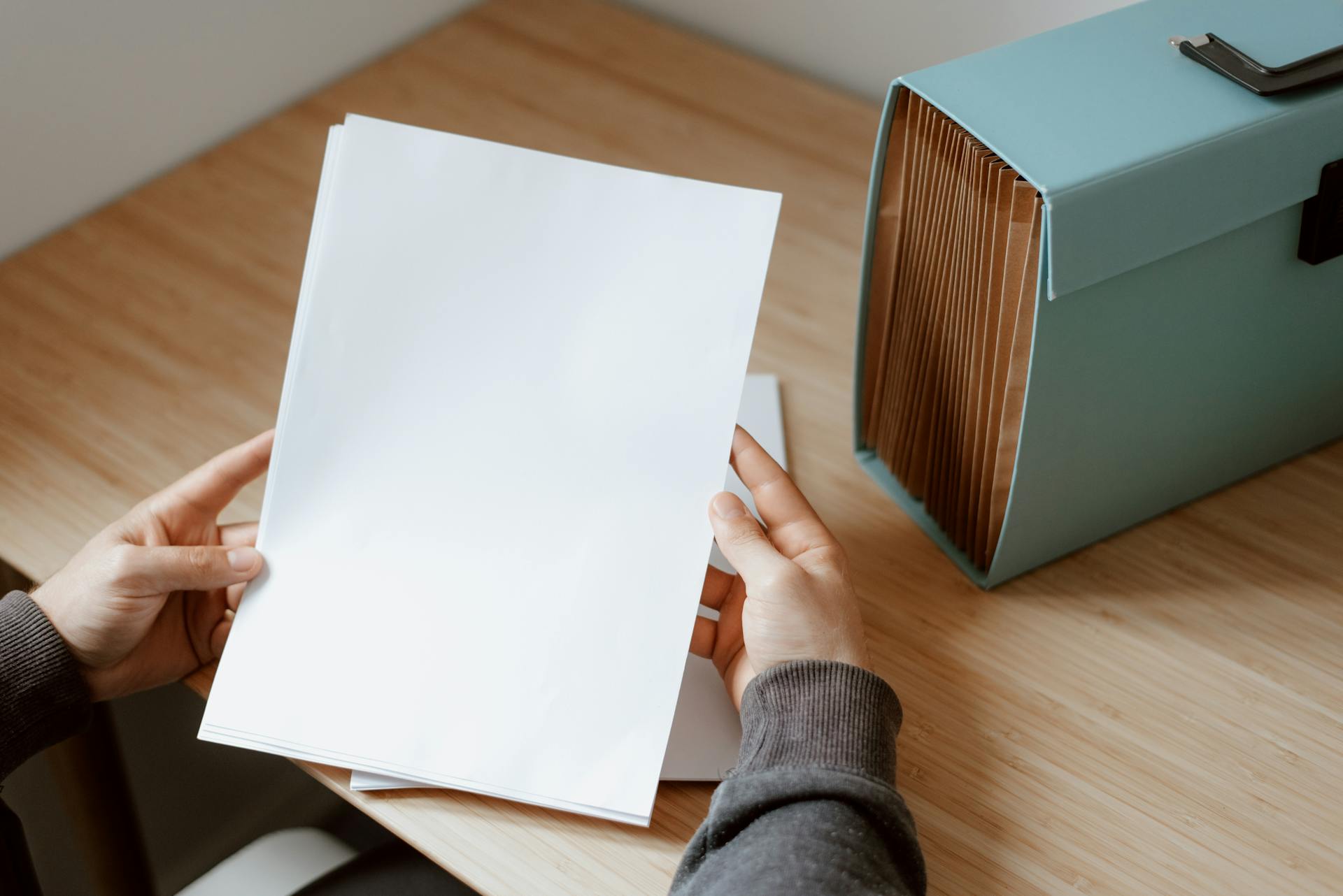 Hands holding a blank sheet of paper next to a turquoise file box on a wooden desk.