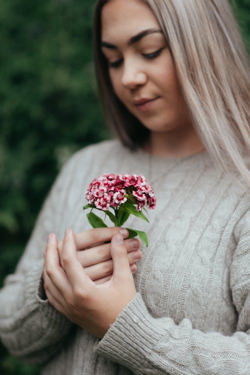 Crop young gentle female in knitted sweater with ornament and bright blossoming flowers looking down on blurred background