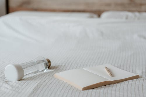 Empty diary with pen near decorative bottle of water on crumpled bed sheet at home