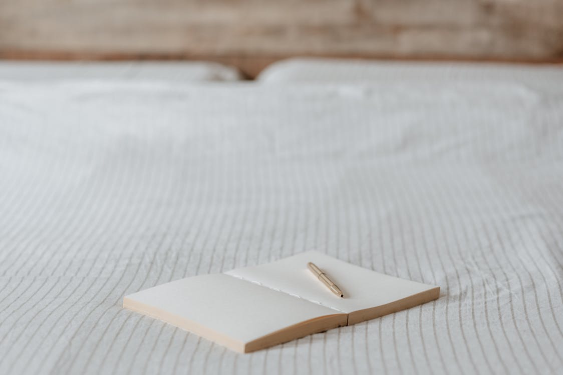 Free Open blank notepad with pen on creased bed sheet with ornament in light bedroom Stock Photo