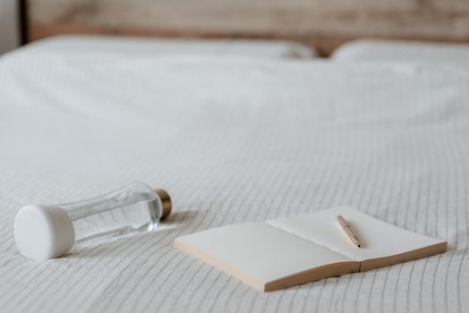 Open empty notepad with pen near small bottle of water on crumpled bed sheet at home
