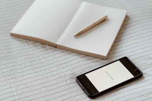Smartphone near empty notebook with pen on bed