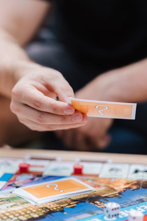 Crop anonymous person with game card playing above board with plastic houses and dices on table