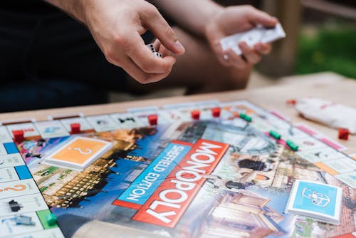 Crop anonymous male player with dices and fake money playing Monopoly at table with deed cards