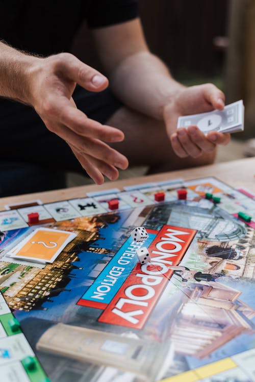 Crop unrecognizable man with fake cash playing board game while rolling dices at table with cards and plastic houses