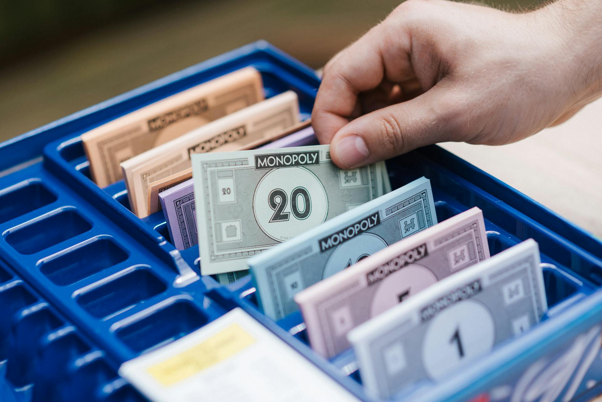 A hand organizing Monopoly game money in a blue plastic tray during gameplay.