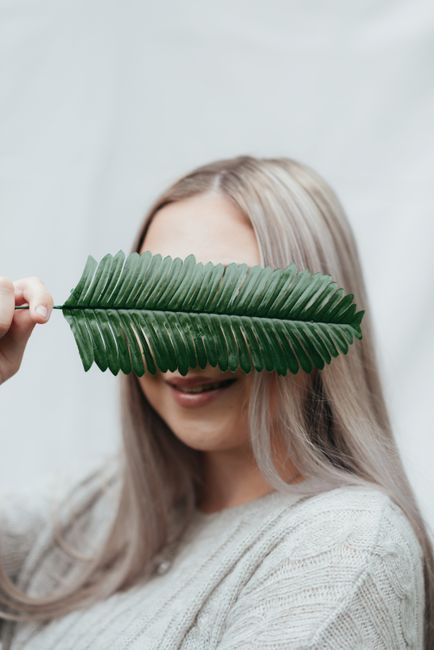Glad woman covering eyes with fresh green leaf of plant while smiling against white background