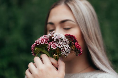 Free Peaceful female with closed eyes smelling bunch of blooming flowers of Turkish carnation while standing against green plants Stock Photo