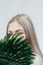 Young female covering half face with green branch of fresh exotic plant against white background
