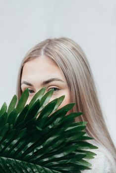 Young female covering half face with green branch of fresh exotic plant against white background