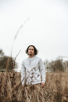 一位表情严肃的亚洲男性站在长满枯草的草地上，从低角度拍摄