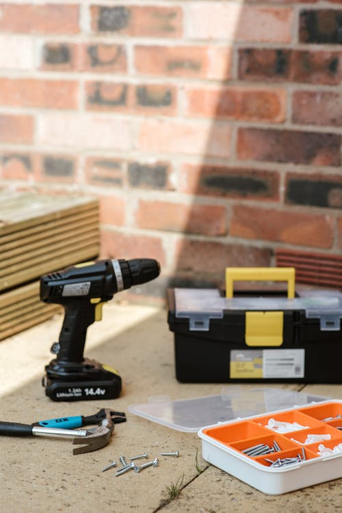 Cordless electric screwdriver placed on floor near boxes with various instruments and hammers against brick wall in sunlight on street