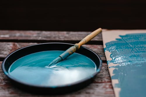 Dirty paintbrush dipped in bowl with azure paint near colorful turquoise carton on wooden bench in street with black background