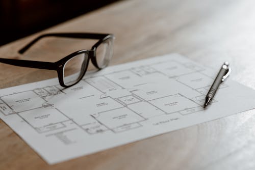 From above of eyeglasses near pen on plan placed on wooden table in apartment in soft daylight