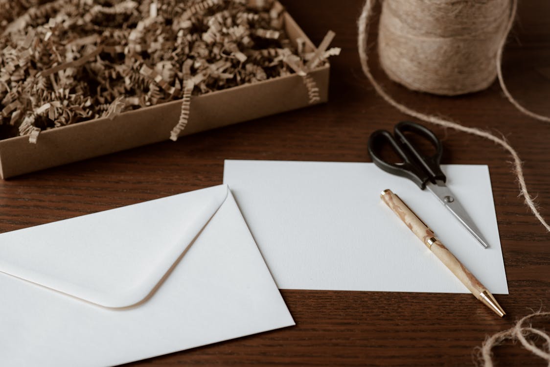 Biała koperta i kartka na stole w otoczeniu nożyczek, długopisu, sznurka i wypełniacza do paczek.