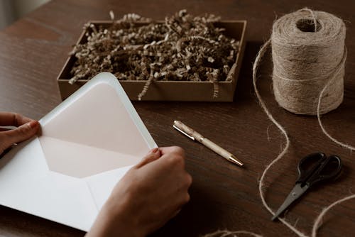 High angle crop anonymous female holding empty envelope above desk with tying twine scissors and box with crinkle paper filler