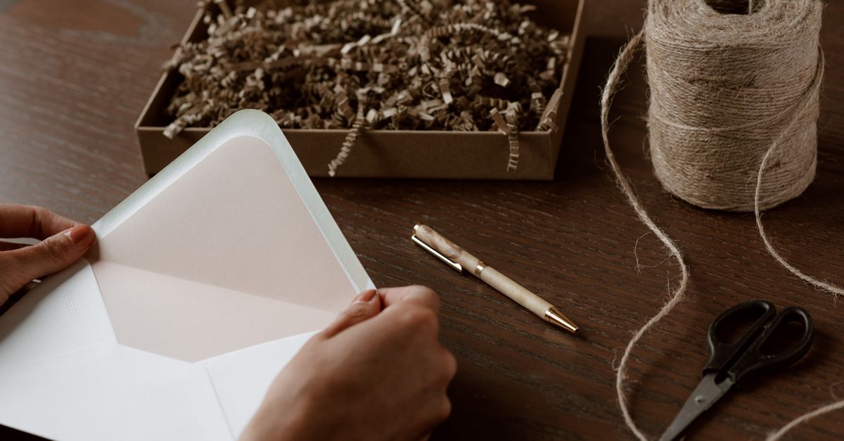 High angle crop anonymous female holding empty envelope above desk with tying twine scissors and box with crinkle paper filler