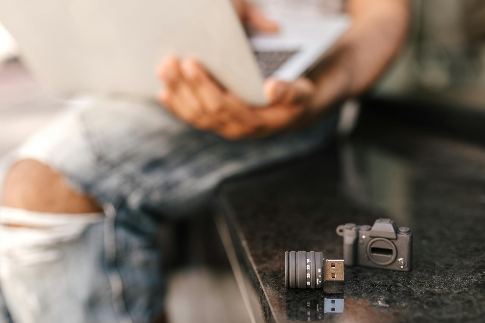 Selective focus of USB stick in shape of camera placed on seat next to crop faceless male freelancer browsing netbook while working remotely