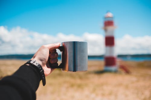 Crop unrecognizable male tourist showing mug of hot drink on ocean coast near beacon under blue cloudy sky