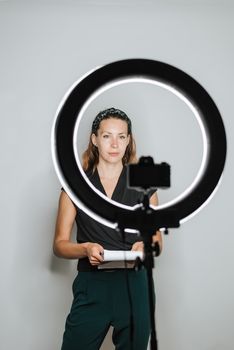 Young female with notebook recording video on photo camera with bright studio lamp on white background