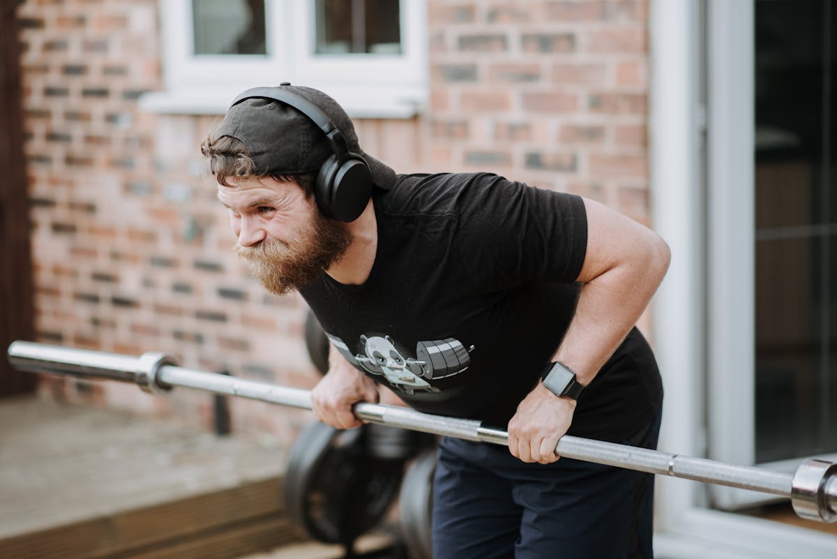 Brutal male with headphones pulling massive metal barbell on blurred background of brick house