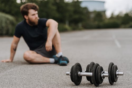 Pensive sporty man sitting on asphalt road with dumbbells