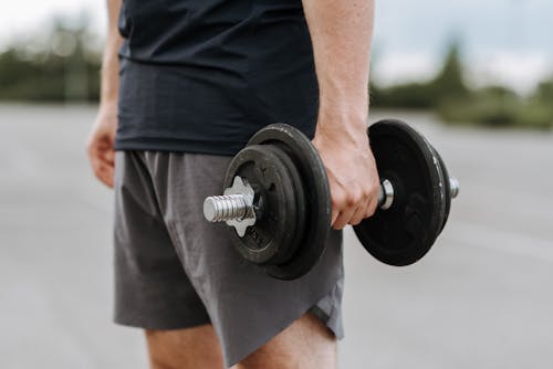 Crop anonymous male in activewear lifting heavy iron dumbbell on blurred background of street