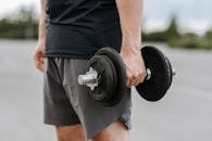 Crop anonymous male in activewear lifting heavy iron dumbbell on blurred background of street