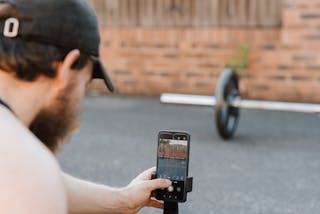 Crop anonymous unshaven sportsman in cap taking photo of barbell on cellphone on pavement in town