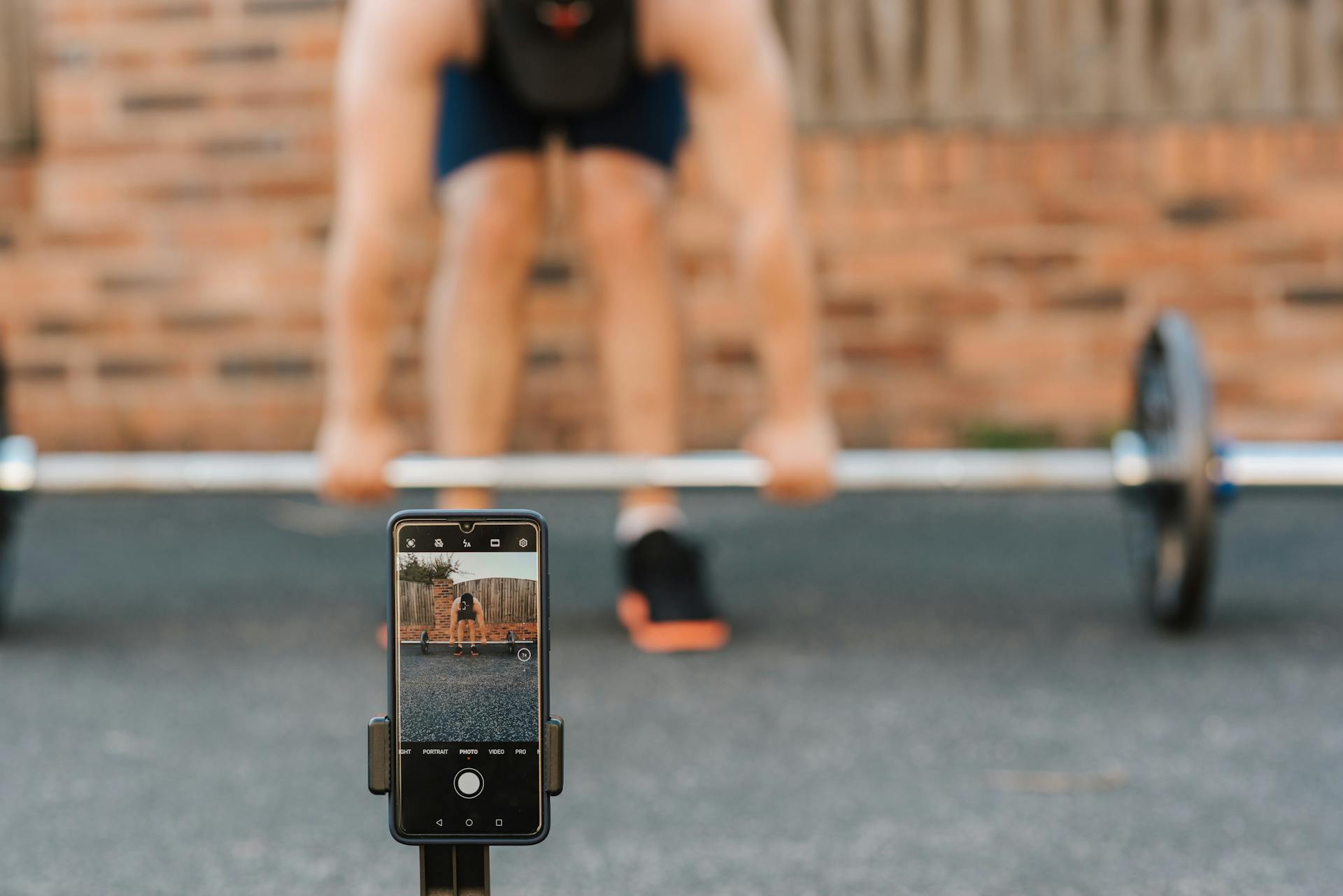 Unrecognizable bodybuilder lifting barbell behind smartphone recording video in town