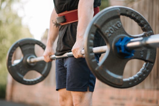 Crop anonymous sportsman in weight belt lifting heavy barbell during training near wall in town