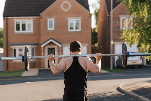 Back view of anonymous fit bodybuilder in cap squatting with barbell while working out on pavement against houses in sunlight
