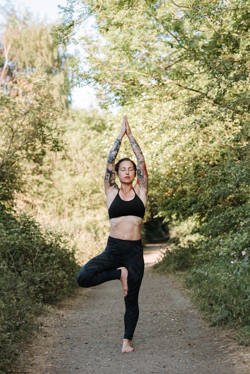 Young reflective tattooed female with closed eyes standing in Vrksasana pose while practicing yoga on walkway between green trees