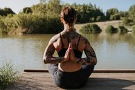 Back view of anonymous tattooed female in sportswear sitting in Reverse Prayer pose on wooden dock against river