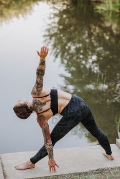 Back view of anonymous flexible barefoot female performing Trikonasana pose while practicing yoga on dock against river