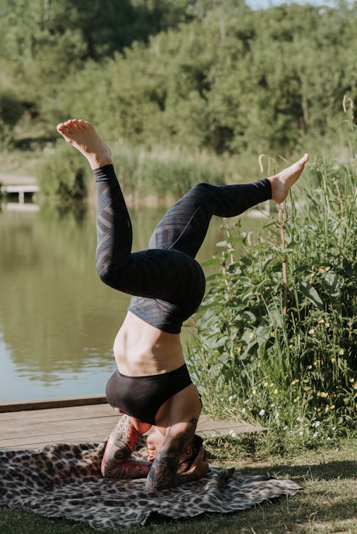 Free Anonymous tattooed female in sportswear balancing on head while practicing yoga on shore against pond Stock Photo