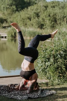 Anonymous tattooed female in sportswear balancing on head while practicing yoga on shore against pond