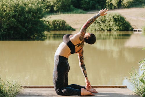 Side view of unrecognizable flexible tattooed lady in sports clothes standing on knees with raised arm on dock against river