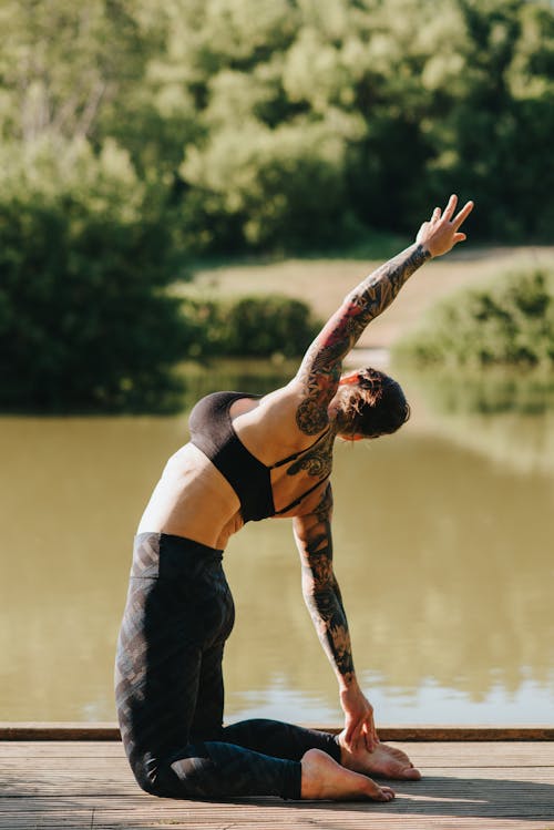 Faceless tattooed woman with raised arm practicing yoga on dock