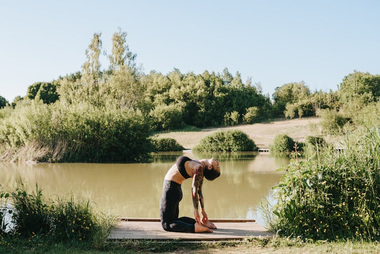 Flexible Woman Performing Camel Pose On Dock Against Lake