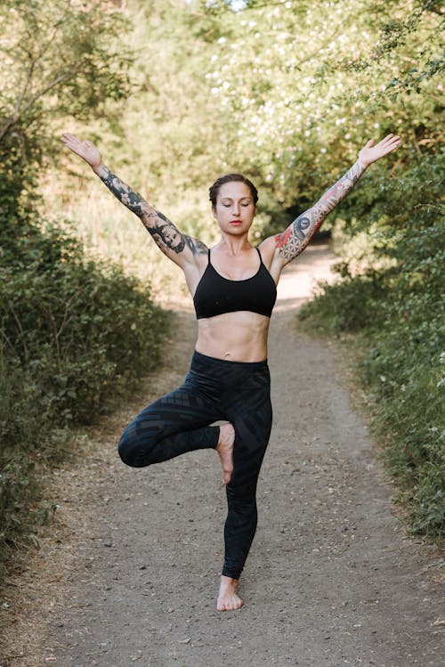 Young mindful tattooed female with closed eyes showing Vrksasana pose with raised arms while practicing yoga on pathway