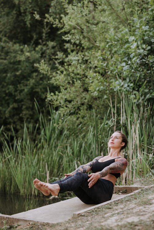 Young attentive tattooed female in sportswear sitting in Navasana pose while practicing yoga on pier and looking up